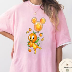 Op de afbeelding: Zachtroze T-shirt met een cartoon sinaasappelpersonage dat sinaasappels en een Mickey Mouse-vormige ballon vasthoudt. Het personage is oranje met groene bladeren en is omringd door sinaasappelschijfjes en kleine sinaasappels. Het shirt heeft een witte kraag.