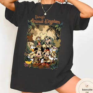 May include: Black t-shirt featuring a graphic of Disney's Animal Kingdom characters, including Mickey and Minnie Mouse, Donald Duck, and Goofy, in safari outfits. The text "Disney's Animal Kingdom" is displayed above the characters.