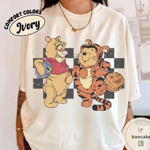 May include: Ivory Comfort Colors t-shirt featuring a vintage-style graphic of two cartoon tigers. One tiger is dressed in a red shirt and holding a blue bag, while the other is in a tiger costume and holding a jack-o'-lantern. A black and white checkered pattern is behind the tigers.