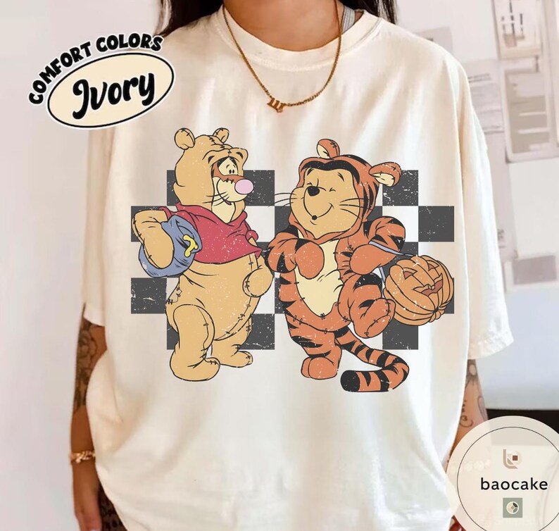 May include: Ivory Comfort Colors t-shirt featuring a vintage-style graphic of two cartoon tigers. One tiger is dressed in a red shirt and holding a blue bag, while the other is in a tiger costume and holding a jack-o'-lantern. A black and white checkered pattern is behind the tigers.