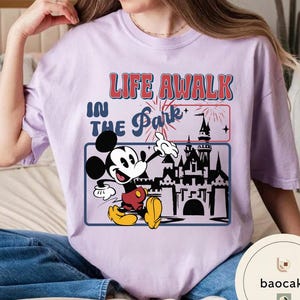 May include: A lilac t-shirt with a graphic of Mickey Mouse and the text "LIFE AWALK IN THE Park". Mickey is depicted in red shorts, yellow shoes, and white gloves. The design includes a castle and fireworks.