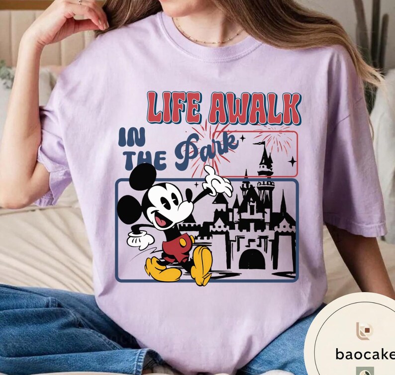 May include: A lilac t-shirt with a graphic of Mickey Mouse and the text "LIFE AWALK IN THE Park". Mickey is depicted in red shorts, yellow shoes, and white gloves. The design includes a castle and fireworks.