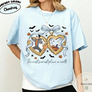 May include: Pale blue t-shirt with a Halloween graphic. The design includes two heart-shaped lockets with cartoon characters, bats, spiders, and pumpkins. The text on the shirt reads "The most scariest place on earth."