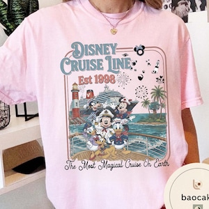 Puede incluir: Camiseta rosa claro con un dise&ntilde;o de Disney Cruise Line. El dise&ntilde;o incluye a Mickey Mouse, Minnie Mouse, Donald Duck y Goofy en un crucero, con el texto "Disney Cruise Line Est 1998" y "The Most Magical Cruise On Earth."