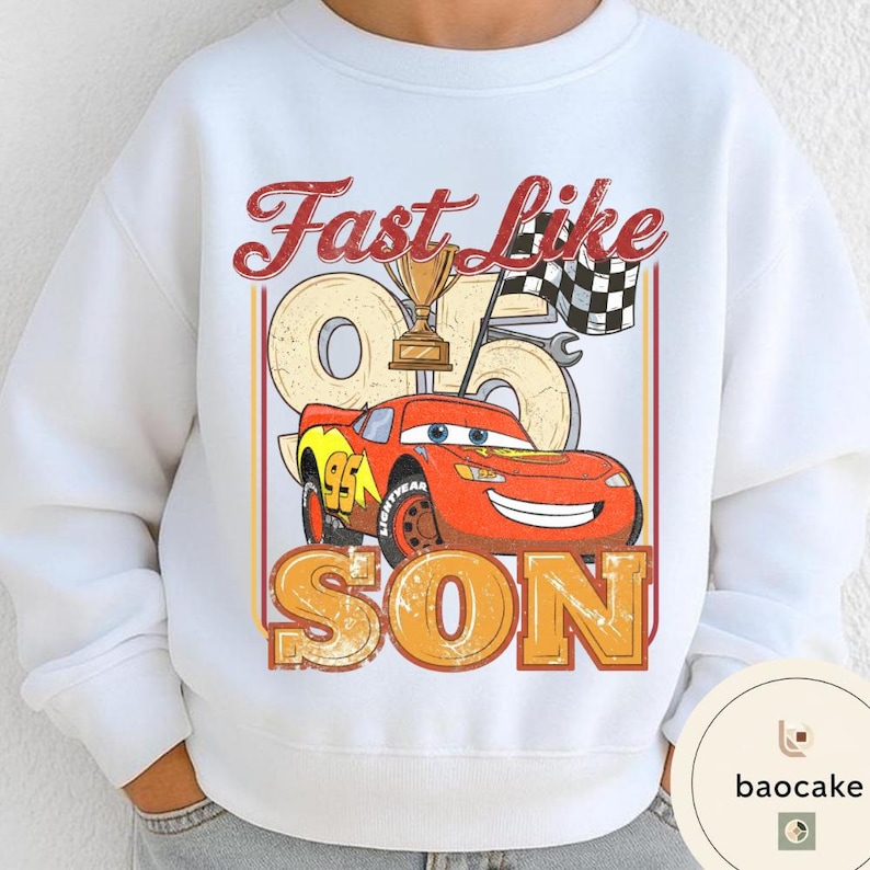 May include: White sweatshirt featuring a graphic of a red race car with the text "Fast Like Son". The design includes a trophy, checkered flag, and the number 95. The sweatshirt is a children's apparel item.