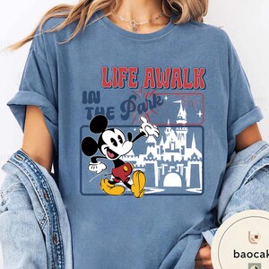 May include: A blue t-shirt with a graphic of Mickey Mouse and the text "LIFE AWALK IN THE Park+". The design includes a castle and fireworks. Mickey is in red shorts and yellow shoes. The shirt is a relaxed, casual fit.