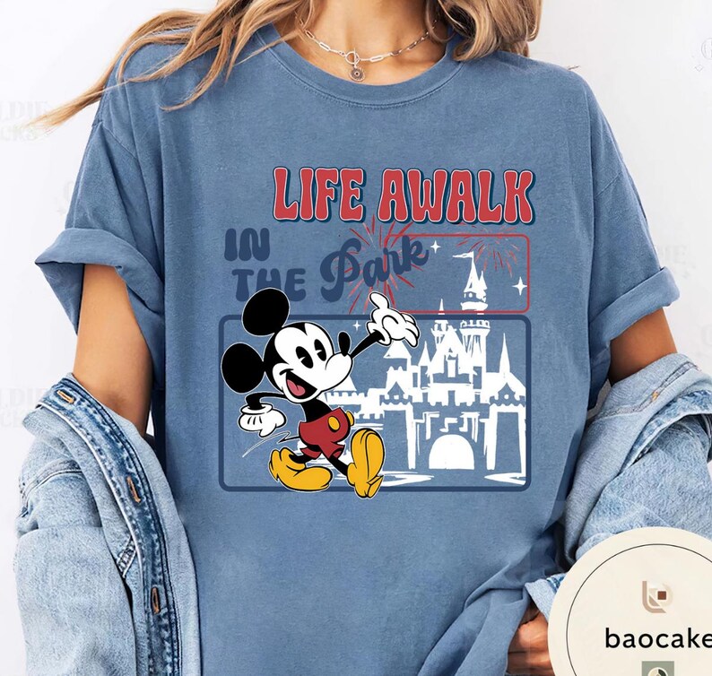 May include: A blue t-shirt with a graphic of Mickey Mouse and the text "LIFE AWALK IN THE Park+". The design includes a castle and fireworks. Mickey is in red shorts and yellow shoes. The shirt is a relaxed, casual fit.