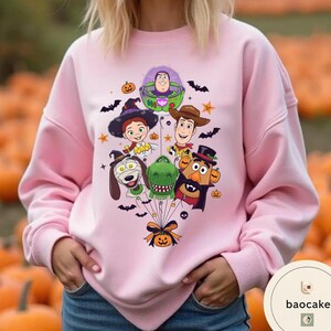 Pode incluir: Uma camisola rosa claro com uma impress&atilde;o gr&aacute;fica de personagens de Toy Story vestidos com fatos de Halloween, incluindo Buzz Lightyear, Woody, Jessie, Rex, Slinky Dog e Mr. Potato Head, todos atados a uma ab&oacute;bora com um la&ccedil;o preto.