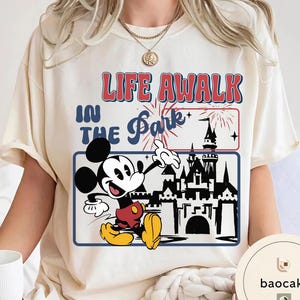May include: Cream-coloured t-shirt with a graphic of Mickey Mouse pointing towards a castle. The text reads "LIFE AWALK IN THE Park". The design includes fireworks and a blue border.