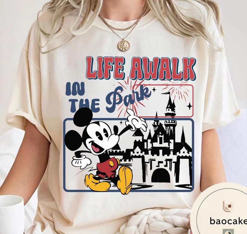 May include: Cream-coloured t-shirt with a graphic of Mickey Mouse pointing towards a castle. The text reads "LIFE AWALK IN THE Park". The design includes fireworks and a blue border.