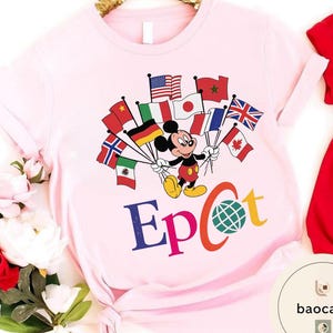 May include: A pale pink t-shirt with a graphic of Mickey Mouse holding flags from different nations. The word "Epcot" is printed in colourful letters below the character, with a globe graphic. The shirt has rolled-up sleeves.