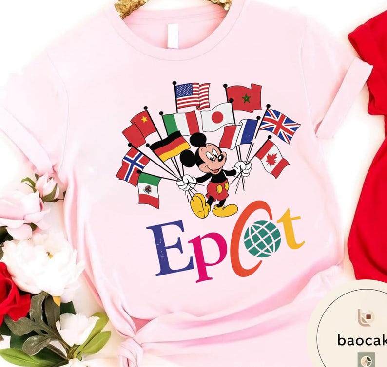 May include: A pale pink t-shirt with a graphic of Mickey Mouse holding flags from different nations. The word "Epcot" is printed in colourful letters below the character, with a globe graphic. The shirt has rolled-up sleeves.