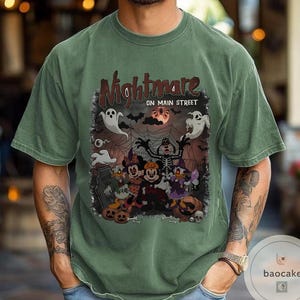 May include: Green t-shirt with a Halloween-themed graphic. The design features the text "Nightmare on Main Street" above cartoon characters, ghosts, bats, and pumpkins. The shirt is a solid green color.
