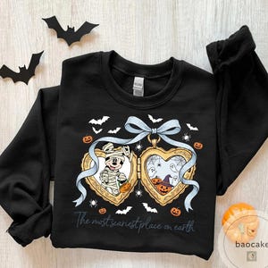 May include: Black sweatshirt with a Halloween design. It features a heart-shaped locket with Mickey Mouse as a mummy and a spooky scene with pumpkins and ghosts. The text reads "The most scariest place on earth".
