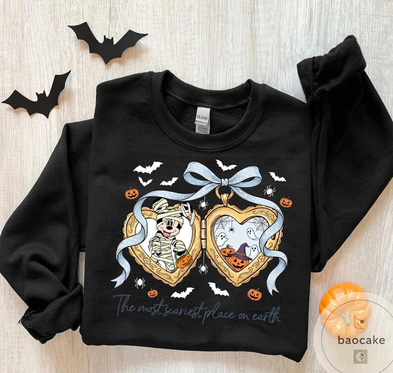 May include: Black sweatshirt with a Halloween design. It features a heart-shaped locket with Mickey Mouse as a mummy and a spooky scene with pumpkins and ghosts. The text reads "The most scariest place on earth".
