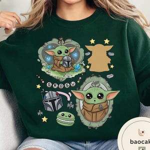 May include: A dark green crewneck sweatshirt featuring a graphic print of Grogu from Star Wars. The design includes Grogu holding a helmet, a silhouette, a portrait in a frame, a silver helmet, a green macaron, and the text GROGU on a banner.