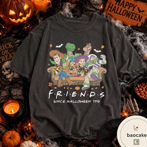 May include: A charcoal grey short-sleeve t-shirt featuring a graphic print of Toy Story characters sitting on a sofa. The text reads FRIENDS SINCE HALLOWEEN 1995. The design includes small pumpkins, bats, and autumn leaves surrounding the characters.