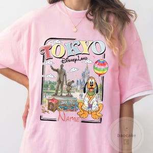 Puede incluir: Camiseta rosa con un gr&aacute;fico que presenta el texto "TOKYO Disneyland" e ilustraciones de personajes de Disney, incluyendo a Pluto, un castillo y un globo arco&iacute;ris. La camiseta tiene cuello blanco.