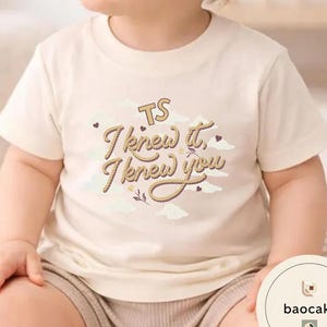 K&ouml;nnte beinhalten: Ein cremefarbenes Kurzarm-T-Shirt mit einem Grafikdruck aus Wolken und dem Text "TS I knew it, I knew you" in einer Schriftart, die wie ein Seil aussieht. Das Design enth&auml;lt kleine Herzakzente und ein florales Element auf der Brust.
