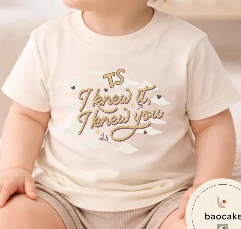 K&ouml;nnte beinhalten: Ein cremefarbenes Kurzarm-T-Shirt mit einem Grafikdruck aus Wolken und dem Text "TS I knew it, I knew you" in einer Schriftart, die wie ein Seil aussieht. Das Design enth&auml;lt kleine Herzakzente und ein florales Element auf der Brust.