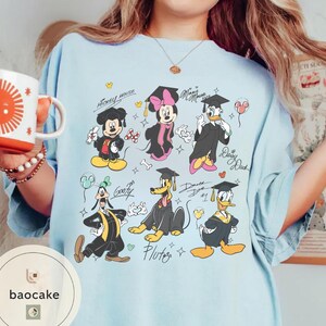 May include: Light blue t-shirt featuring cartoon characters Mickey Mouse, Minnie Mouse, Donald Duck, Daisy Duck, Goofy, and Pluto in graduation attire. The shirt has the text "Mickey Mouse" and "Minnie Mouse".