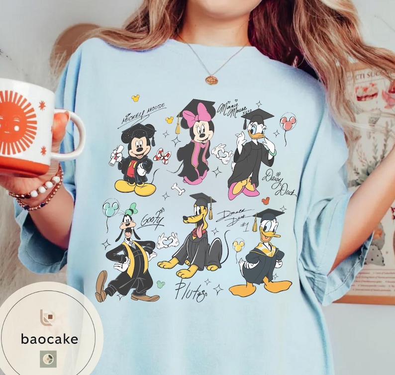 May include: Light blue t-shirt featuring cartoon characters Mickey Mouse, Minnie Mouse, Donald Duck, Daisy Duck, Goofy, and Pluto in graduation attire. The shirt has the text "Mickey Mouse" and "Minnie Mouse".