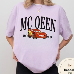 May include: A light purple t-shirt featuring the text "MC QUEEN" in large black letters above a cartoon image of a red race car. The numbers "20" and "26" flank the car. The shirt is a casual style.