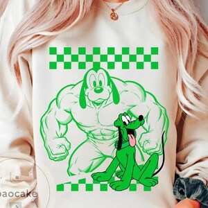 May include: Cream-colored sweatshirt featuring a green cartoon dog design. The design includes a muscular dog figure and a smaller, sitting dog with its tongue out. A green and white checkered pattern is at the top and bottom of the design.