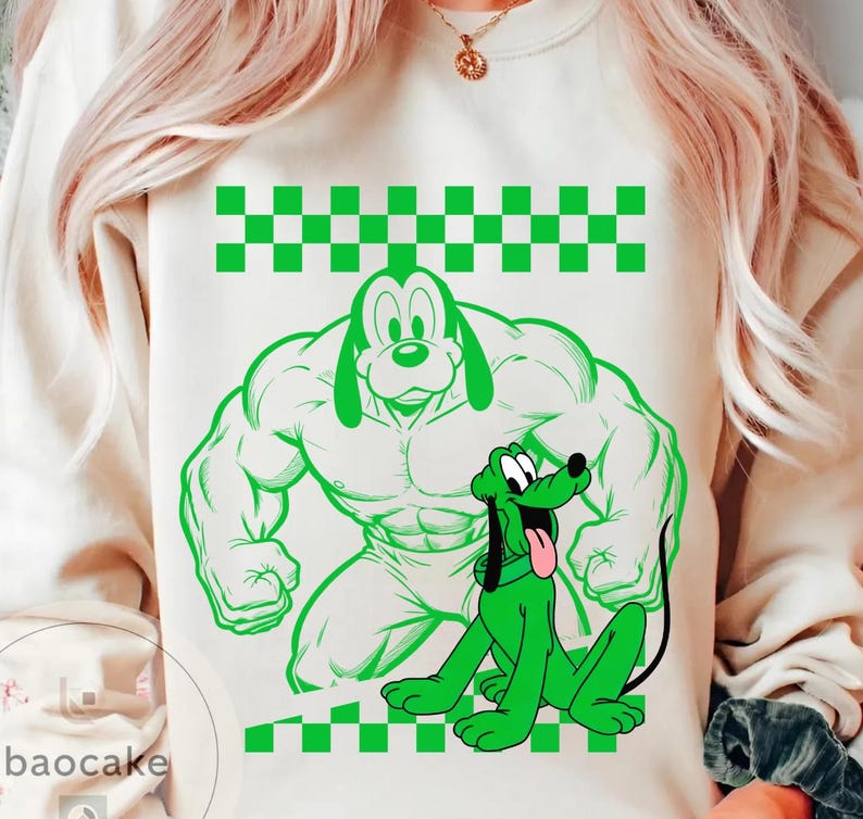 May include: Cream-colored sweatshirt featuring a green cartoon dog design. The design includes a muscular dog figure and a smaller, sitting dog with its tongue out. A green and white checkered pattern is at the top and bottom of the design.