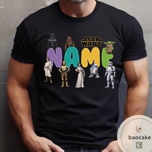 May include: Black t-shirt featuring Star Wars characters and the word "NAME" in colorful bubble letters. The Star Wars logo is in gold. Characters include Darth Vader, Chewbacca, Yoda, and others. A fun, personalized design.