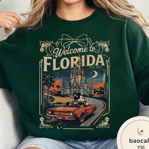 May include: Dark green sweatshirt with a graphic that says "Welcome to Florida" and features a vintage-style illustration of a car with Mickey Mouse driving past a castle. The sweatshirt is a solid color.