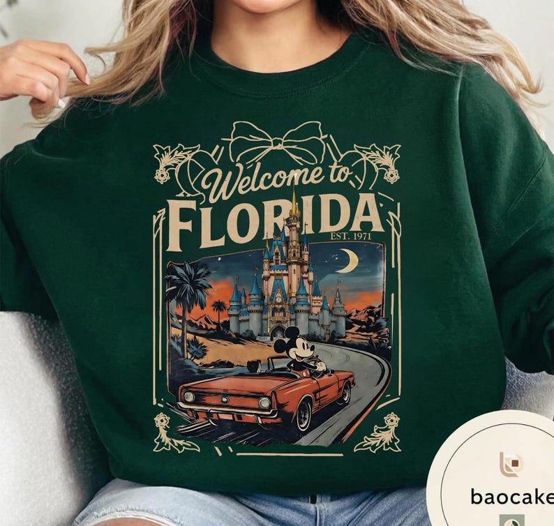 May include: Dark green sweatshirt with a graphic that says "Welcome to Florida" and features a vintage-style illustration of a car with Mickey Mouse driving past a castle. The sweatshirt is a solid color.