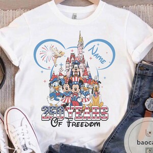 Peut inclure: T-shirt blanc avec un graphisme color&eacute; de personnages Disney en tenues patriotiques, avec un ch&acirc;teau en arri&egrave;re-plan. Le design comprend le texte "250 YEARS OF FREEDOM" et un espace pour un nom, parfait pour une c&eacute;l&eacute;bration th&eacute;matique.