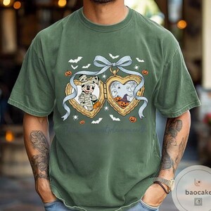 May include: Green t-shirt with a heart-shaped locket design, Halloween themed. The locket shows Mickey Mouse as a mummy and a spooky scene with ghosts and pumpkins. Bats and spiders are also included in the design.