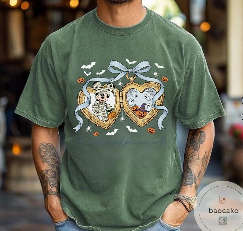 May include: Green t-shirt with a heart-shaped locket design, Halloween themed. The locket shows Mickey Mouse as a mummy and a spooky scene with ghosts and pumpkins. Bats and spiders are also included in the design.