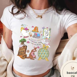 May include: White t-shirt with a colorful graphic featuring cartoon characters and the text "Happiest Mom on Earth." The design includes a teapot, dog, elephant, and kangaroo. The shirt is made of a soft material.