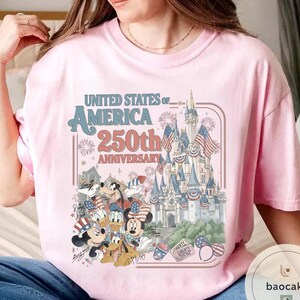 May include: Light pink t-shirt featuring a graphic celebrating the 250th anniversary of the United States of America. The design includes a castle, fireworks, and cartoon characters in patriotic attire.