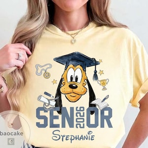 May include: Yellow t-shirt featuring a graduation-themed graphic of Pluto wearing a mortarboard. The design includes the text "SENIOR 2026" and the name "Stephanie". Additional elements include a diploma, stars, and a trophy.