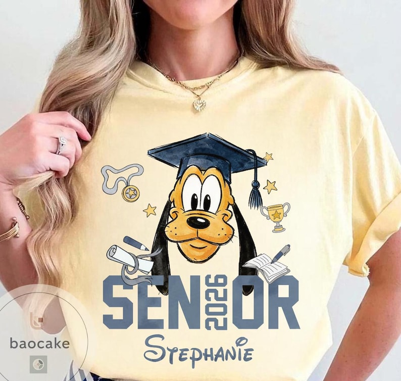 May include: Yellow t-shirt featuring a graduation-themed graphic of Pluto wearing a mortarboard. The design includes the text "SENIOR 2026" and the name "Stephanie". Additional elements include a diploma, stars, and a trophy.