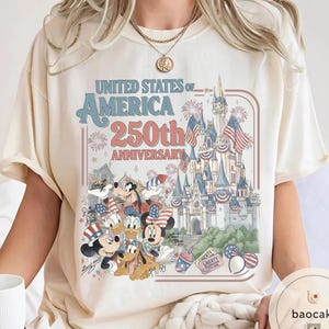 May include: Cream-colored t-shirt featuring a graphic celebrating the United States of America's 250th Anniversary. The design includes a castle, fireworks, and cartoon characters like Mickey and Minnie Mouse, Donald Duck, and Goofy.