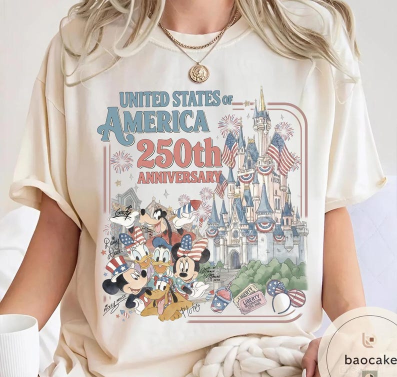 May include: Cream-colored t-shirt featuring a graphic celebrating the United States of America's 250th Anniversary. The design includes a castle, fireworks, and cartoon characters like Mickey and Minnie Mouse, Donald Duck, and Goofy.