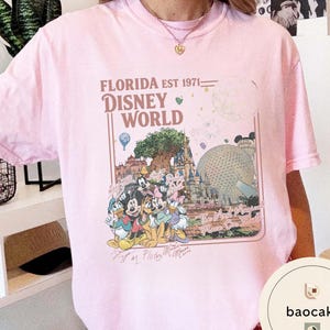 May include: A light pink short-sleeve t-shirt featuring a graphic print of Disney World landmarks and characters. The text reads FLORIDA EST 1971, DISNEY WORLD, and The Most Magical Place On Earth, accompanied by illustrations of Mickey, Minnie, and friends.