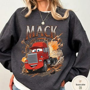 May include: Dark grey sweatshirt with a cartoon illustration of a red semi-truck, the name "MACK" above. The truck has a smiling face and is surrounded by flames and sparks. The sweatshirt is a crew neck style, suitable for casual wear.