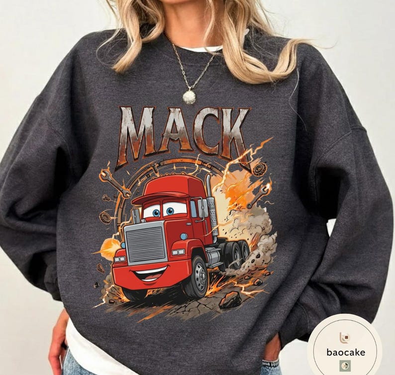 May include: Dark grey sweatshirt with a cartoon illustration of a red semi-truck, the name "MACK" above. The truck has a smiling face and is surrounded by flames and sparks. The sweatshirt is a crew neck style, suitable for casual wear.