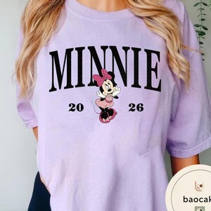 May include: A light purple t-shirt featuring the word "MINNIE" in large black letters above a cartoon image of Minnie Mouse. The numbers "20" and "26" are on either side of the Minnie Mouse graphic. The shirt is a solid color.