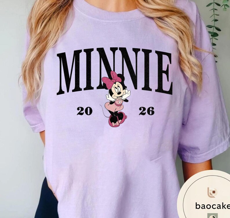 May include: A light purple t-shirt featuring the word "MINNIE" in large black letters above a cartoon image of Minnie Mouse. The numbers "20" and "26" are on either side of the Minnie Mouse graphic. The shirt is a solid color.