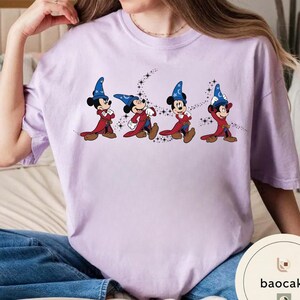 May include: A light purple t-shirt featuring a graphic of Mickey Mouse in a sorcerer's outfit, with a blue hat and red robe. The design shows Mickey in four different poses, with a starry trail.