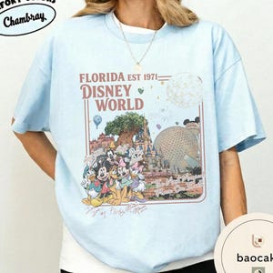 May include: A light blue t-shirt featuring a graphic print with the text FLORIDA EST 1971 DISNEY WORLD and The Most Magical Place On Earth. The design includes illustrations of Disney characters, the Tree of Life, and the Epcot geodesic sphere.