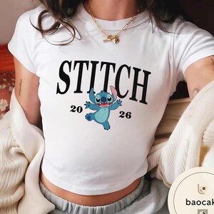 May include: White t-shirt featuring the word "STITCH" in large black letters above a cartoon image of Stitch, the blue alien character. The numbers "20" and "26" are on either side of the character. The shirt is a crew neck style.