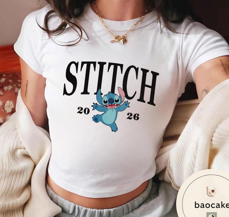 May include: White t-shirt featuring the word "STITCH" in large black letters above a cartoon image of Stitch, the blue alien character. The numbers "20" and "26" are on either side of the character. The shirt is a crew neck style.